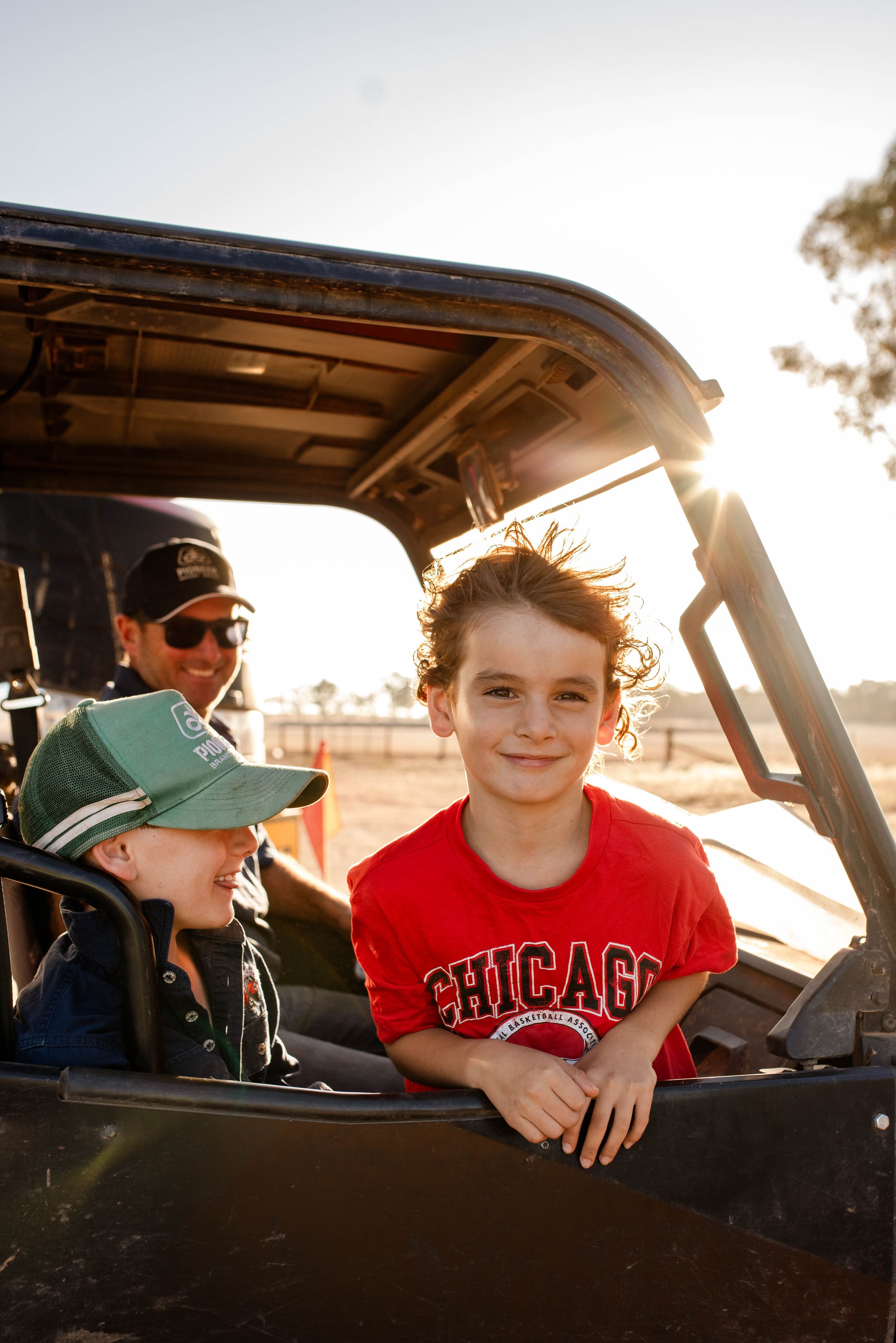 Three children smile inside a UTV on a rural farm at golden hour, the eldest wearing a red Chicago t-shirt.