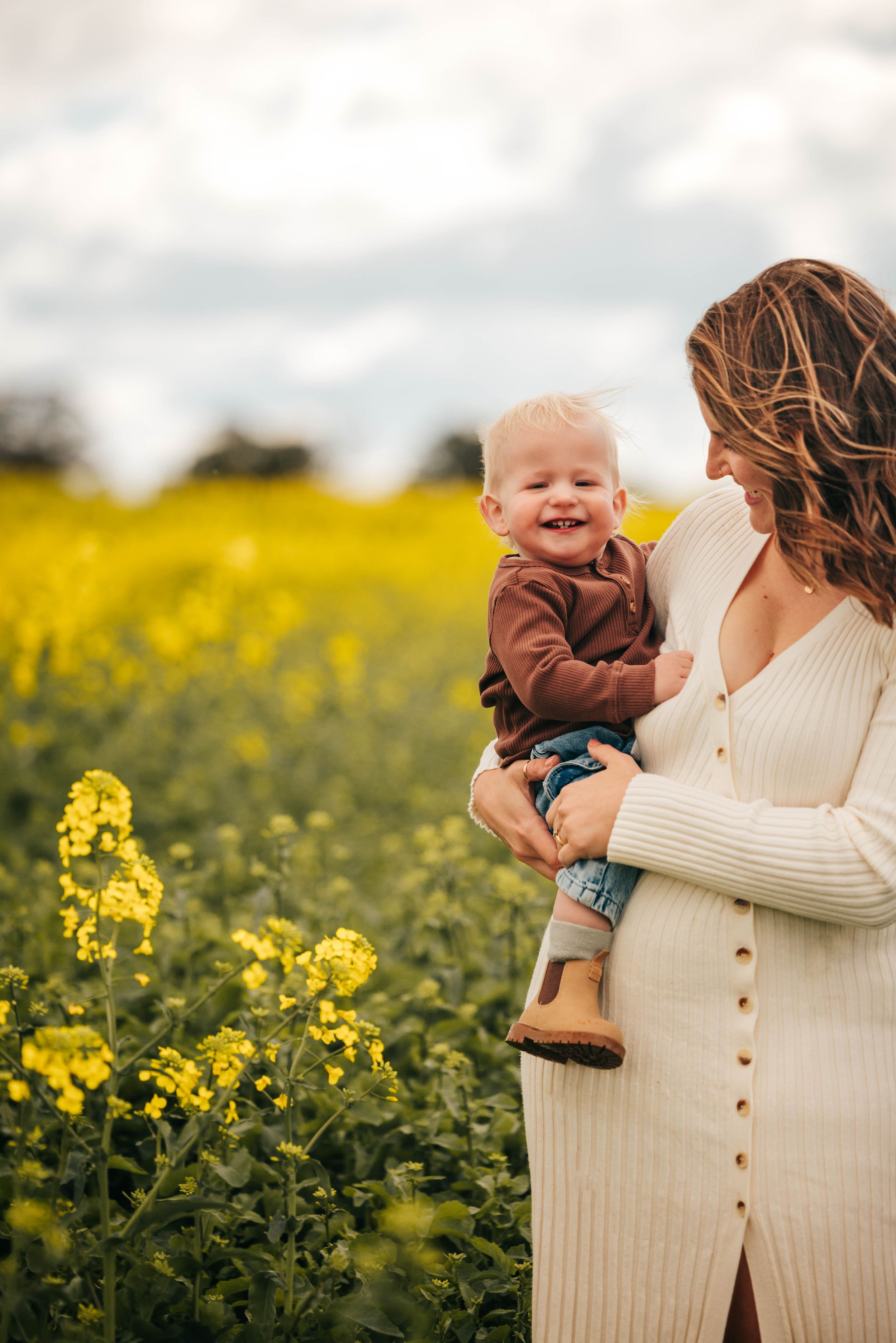 A smiling baby held by a pregnant woman in a white dress standing in a vibrant yellow rapeseed field.