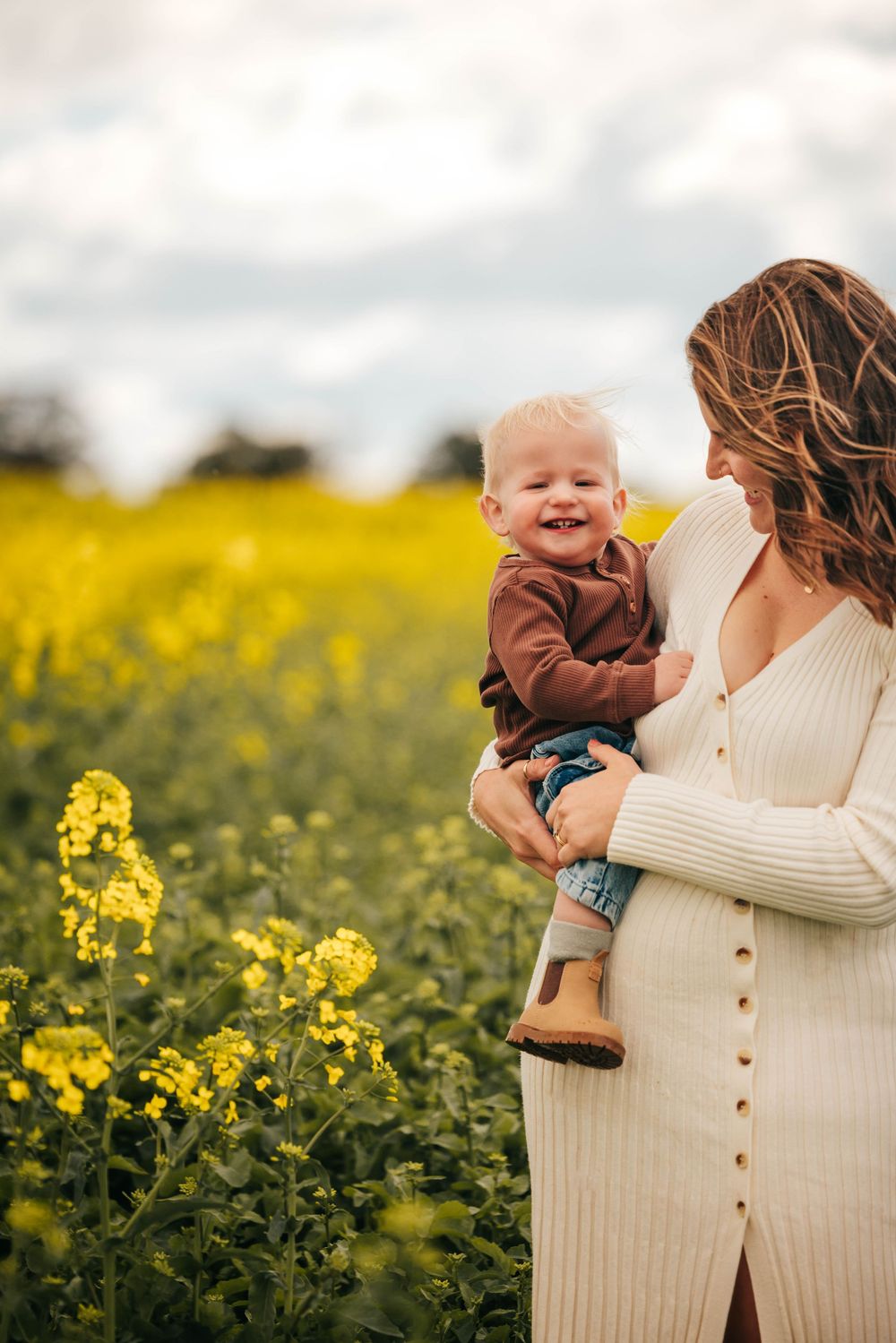 A smiling baby held by a pregnant woman in a white dress standing in a vibrant yellow rapeseed field.