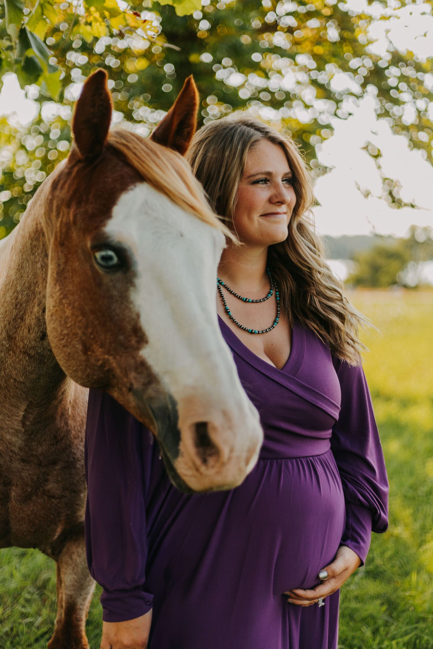 A pregnant woman in a purple dress stands with a brown and white horse in a sunny outdoor field.
