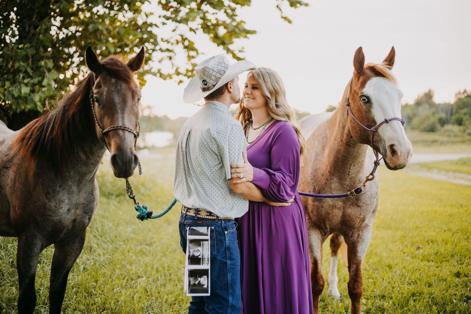A couple in a purple dress and blue shirt poses with their horses in a sunlit field at sunset.