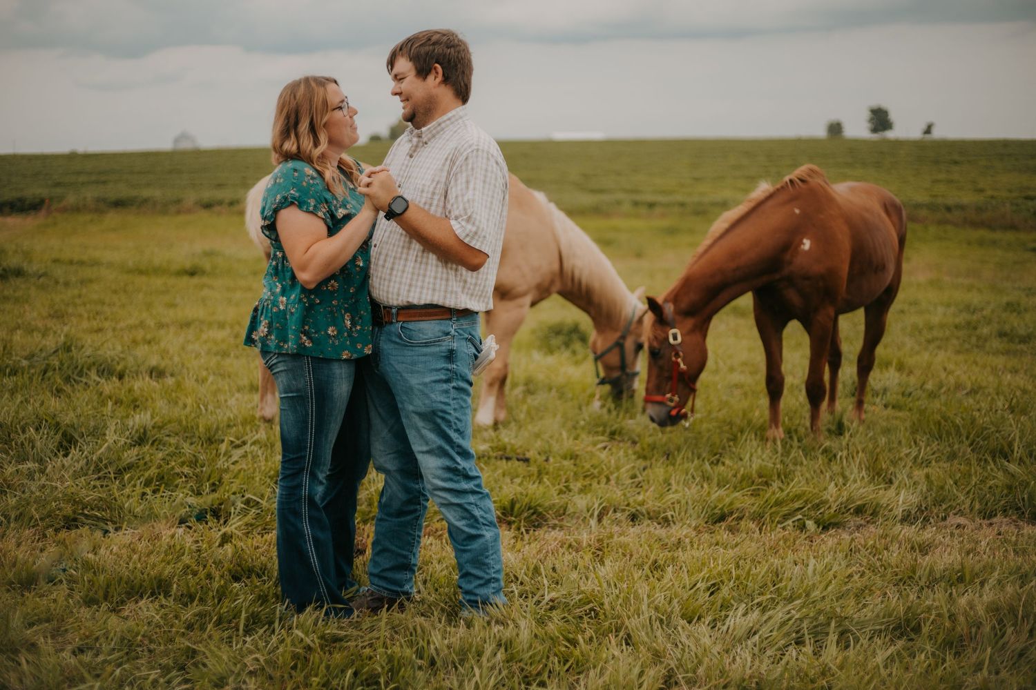 A couple embraces in a field while a brown horse grazes in the background.