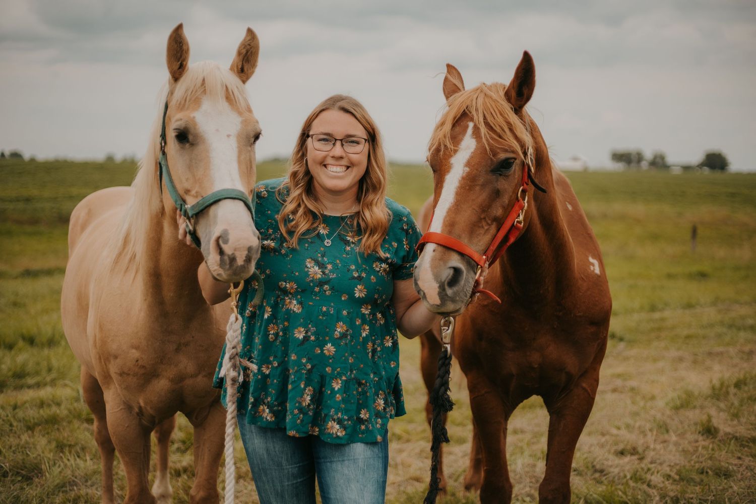 A person in a green floral dress stands between two horses in a pastoral setting.