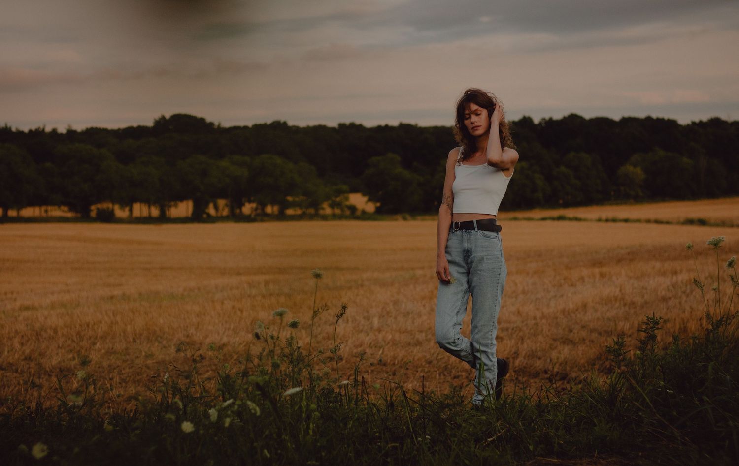 Model posing in farmer's field during the fall. Model is wearing blue jeans and a white tank top.