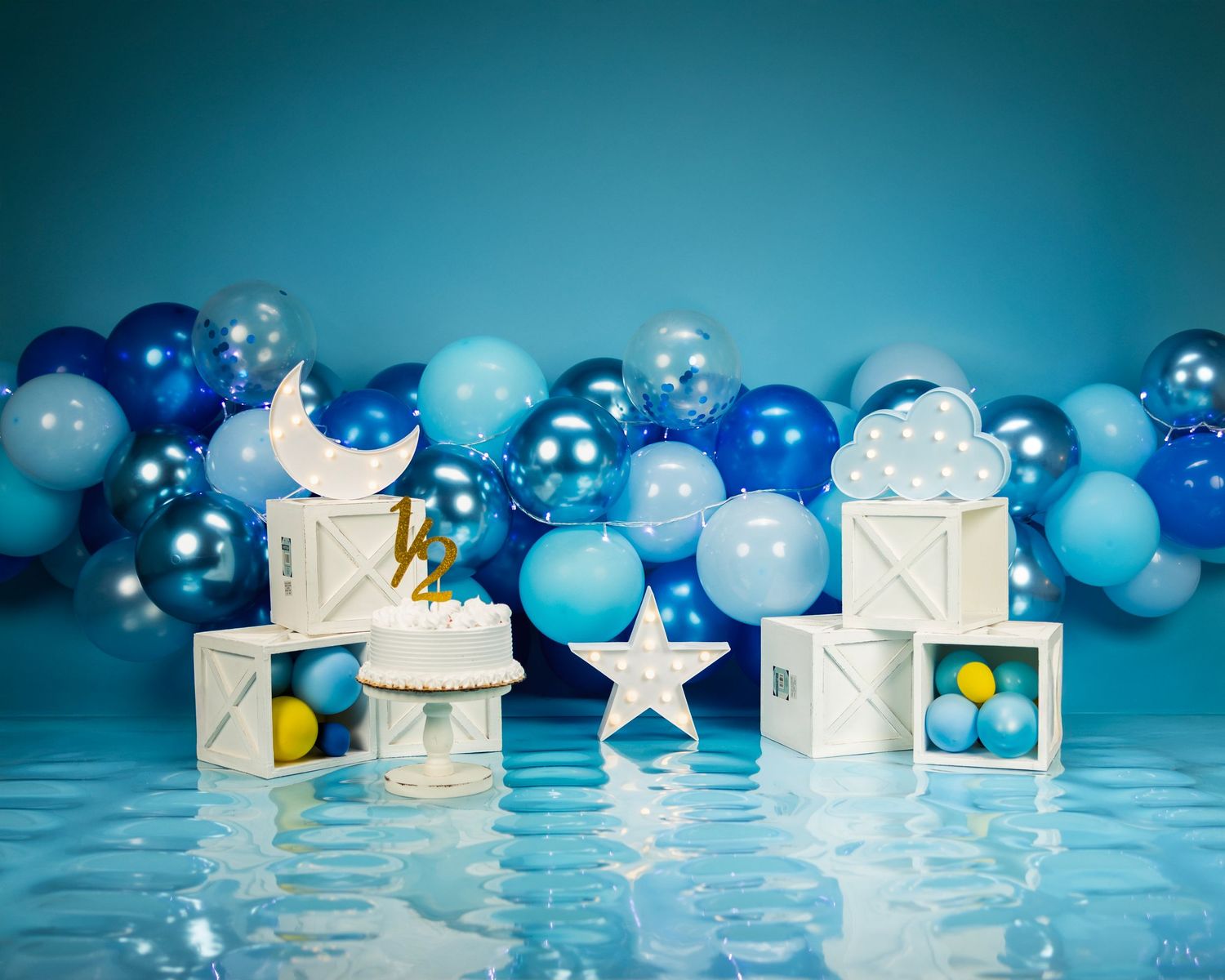 Decorative white gift boxes and glowing blue balloon garland arranged on reflective surface against turquoise backdrop.
