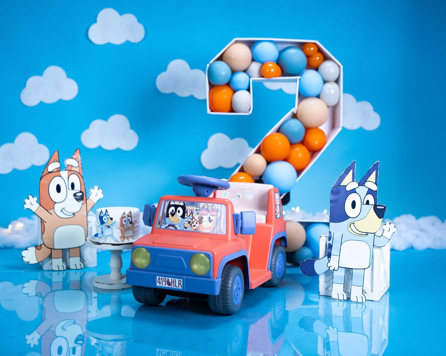 Whimsical toy scene featuring Bluey characters with a red toy truck and number 2 balloon against a blue cloudy backdrop.