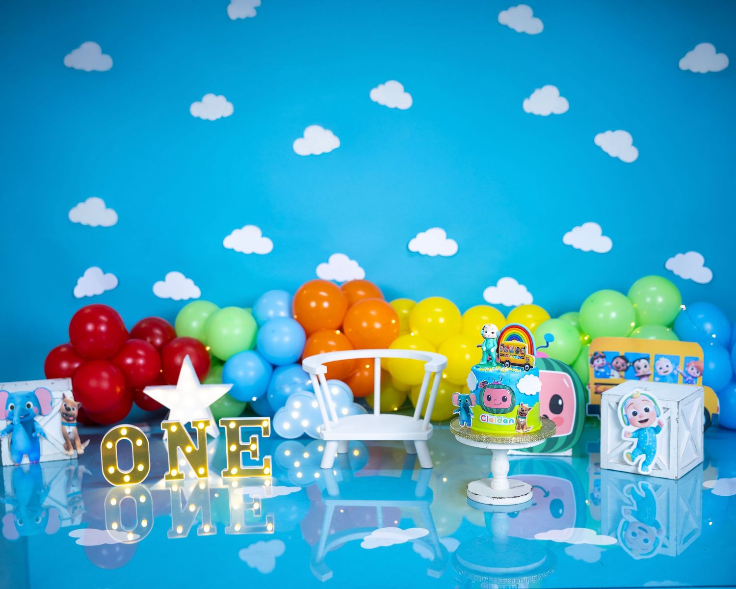 Colorful party decoration setup with blue sky backdrop, balloons, 'ONE' text and baby shower elements on ice-like surfaces.