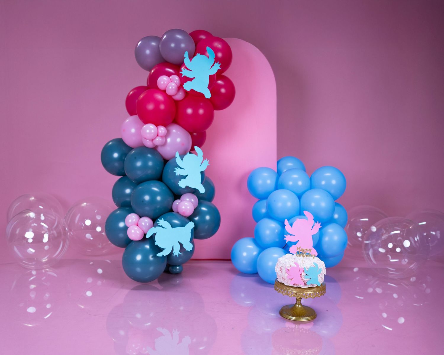 Balloon arrangement in shades of pink, blue and purple with small decorative elements against pink backdrop.