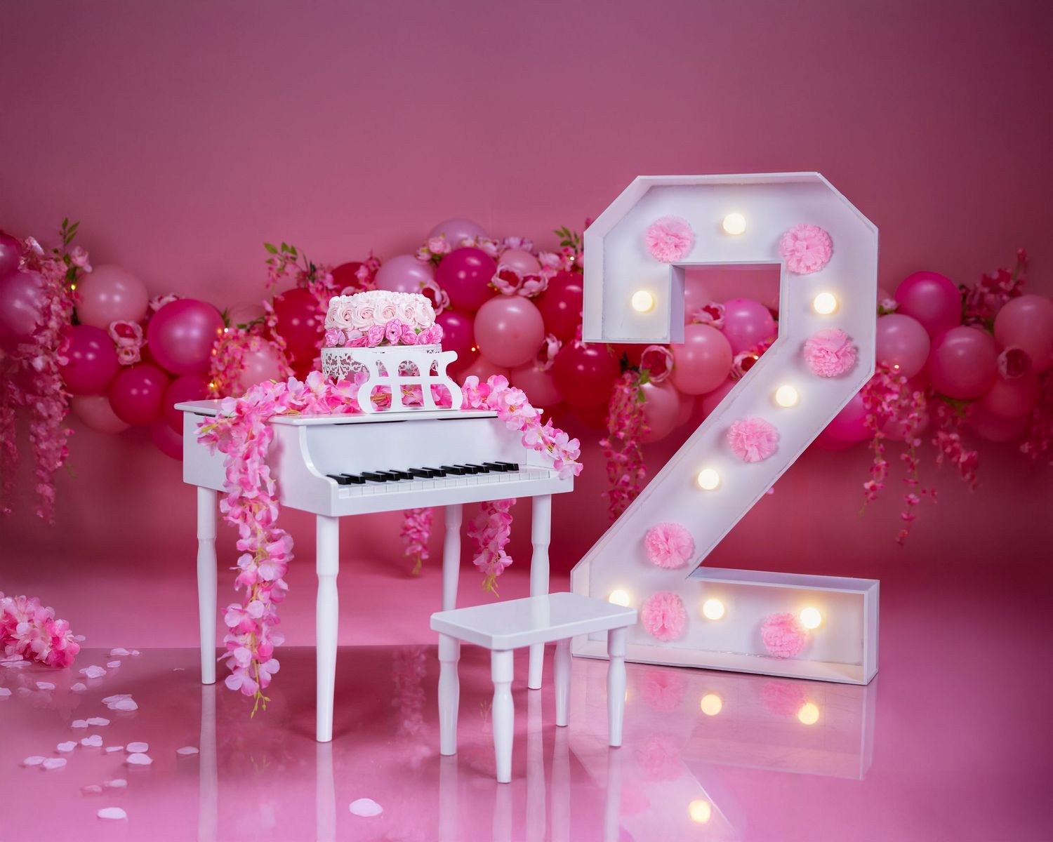 Pink birthday setup with illuminated number 2 marquee light and white piano against pink balloon background.