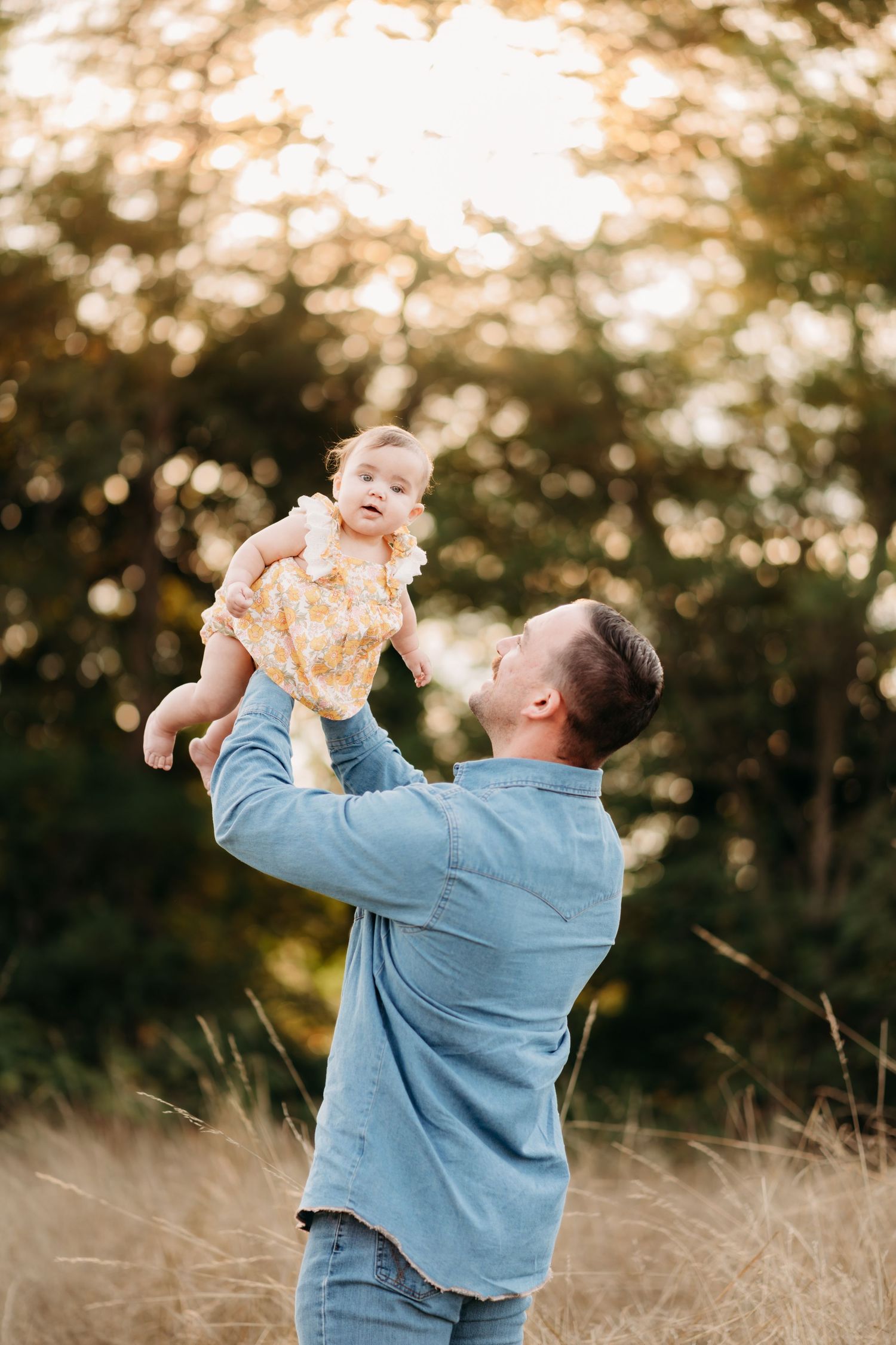 Dad in a blue sweater lifts and plays with a small child in a sunny outdoor field at golden hour at their family photo session in Tacoma, WA