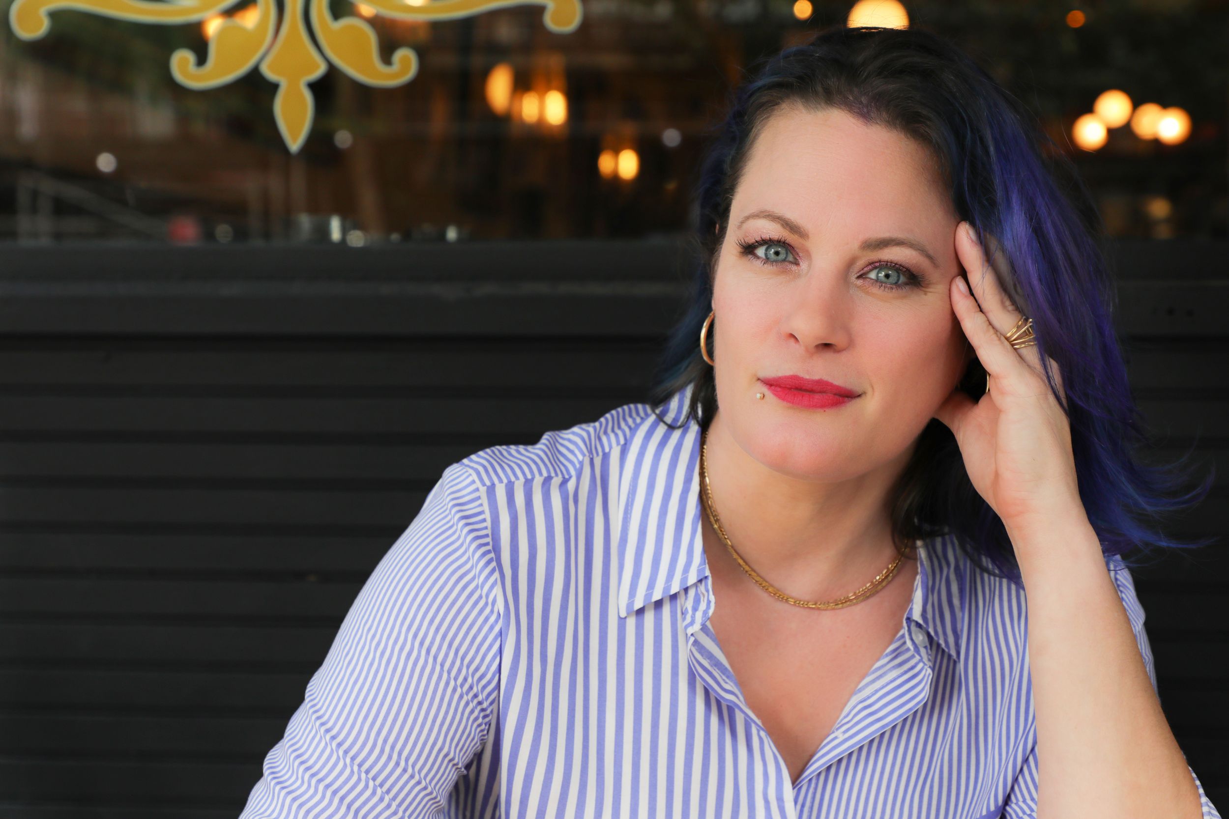 A thoughtful portrait features purple-dyed hair and a striped blue and white button down shirt in a restaurant setting.