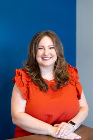 Professional headshot of someone in a bright red ruffled top smiling warmly against a blue background.