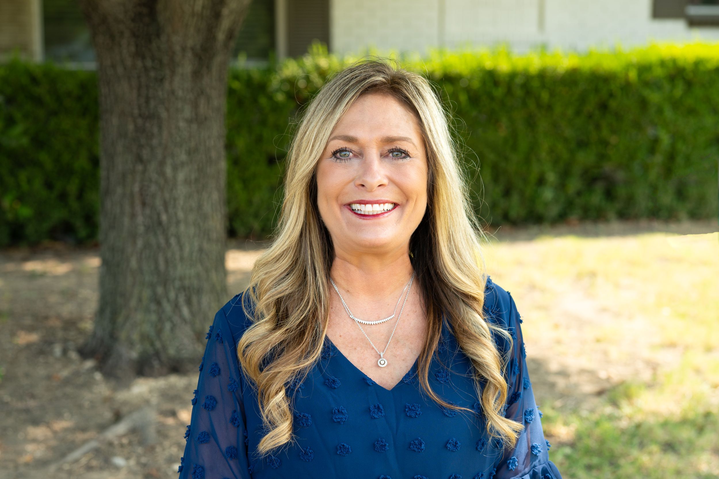 Professional outdoor portrait of someone in a navy blue v-neck top smiling warmly against a garden background.