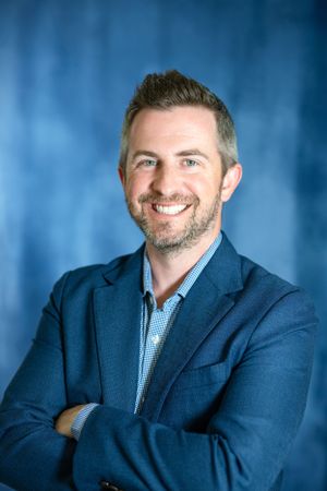 Professional headshot portrait of someone wearing a navy blazer and light blue dress shirt against a blue background.