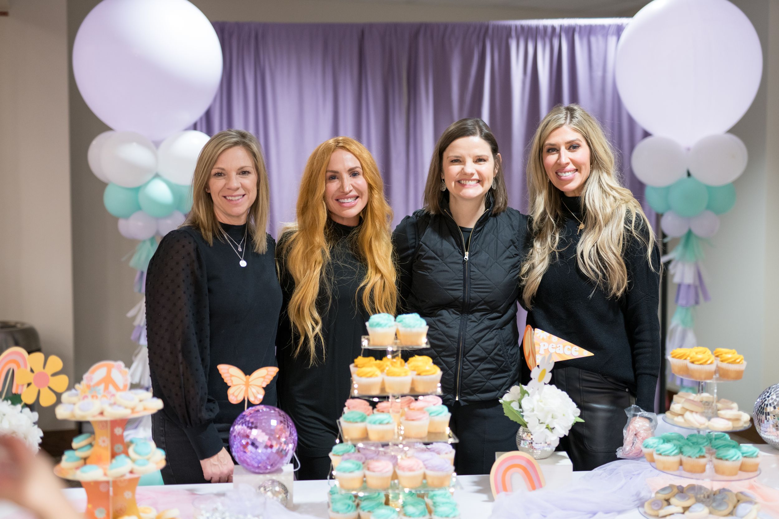 Four women in black outfits smile behind a colorful dessert table with cupcakes and balloon backdrop.