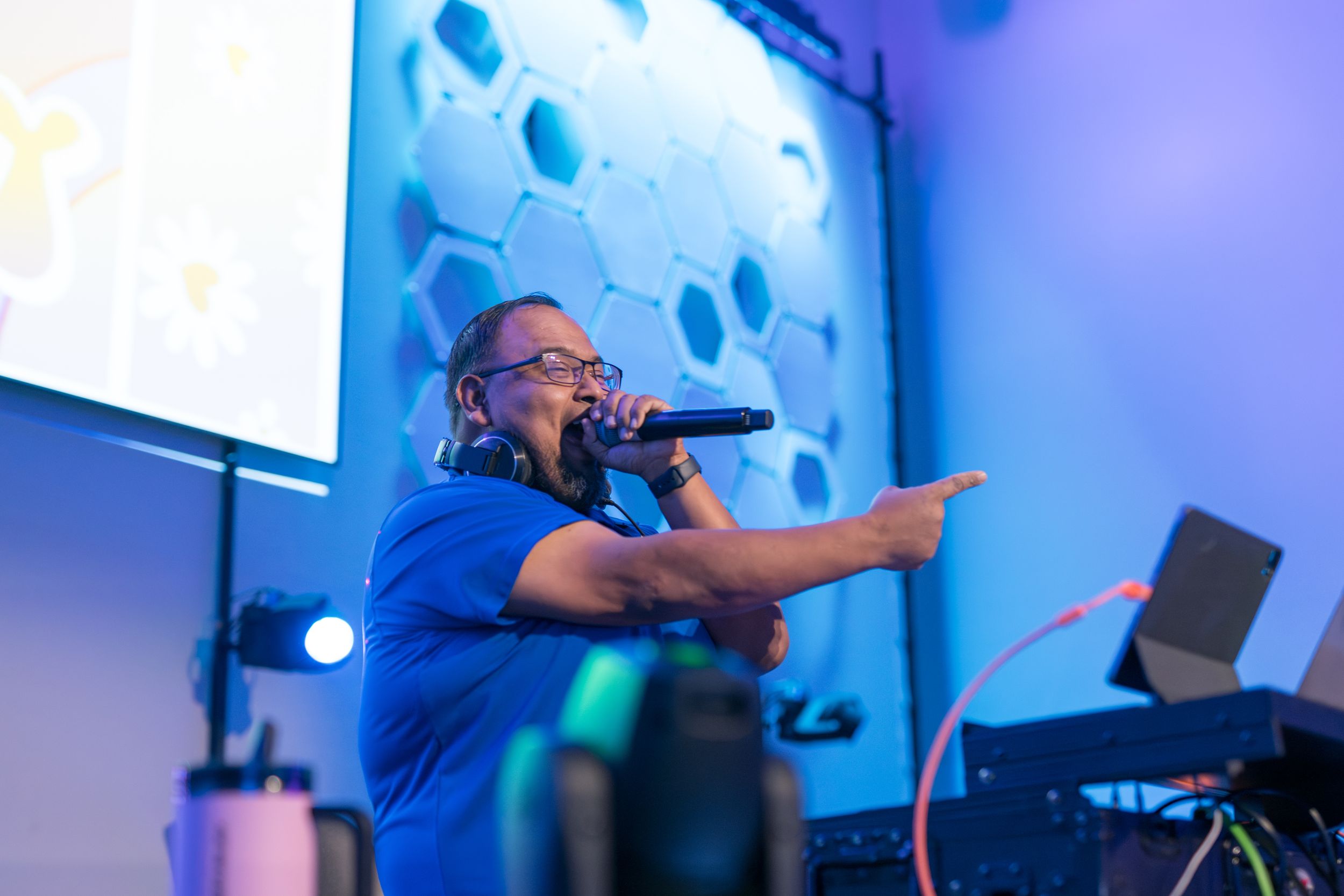 A vocalist in a bright blue shirt performs on stage with electronic equipment and lighting effects in the background.