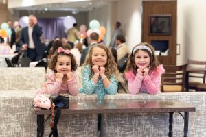 Three excited children lean on counter at a party with balloons and decorations in the background.