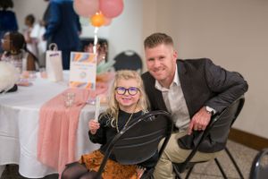 Two people pose together at a party with pink decorations and balloons in a banquet room setting.