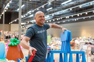 A person wearing a gray Rock Fit t-shirt smiles while holding up blue chairs in a large exhibition hall.