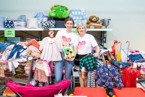Two individuals stand among racks of colorful children's clothing and accessories in a resale or consignment shop setting.
