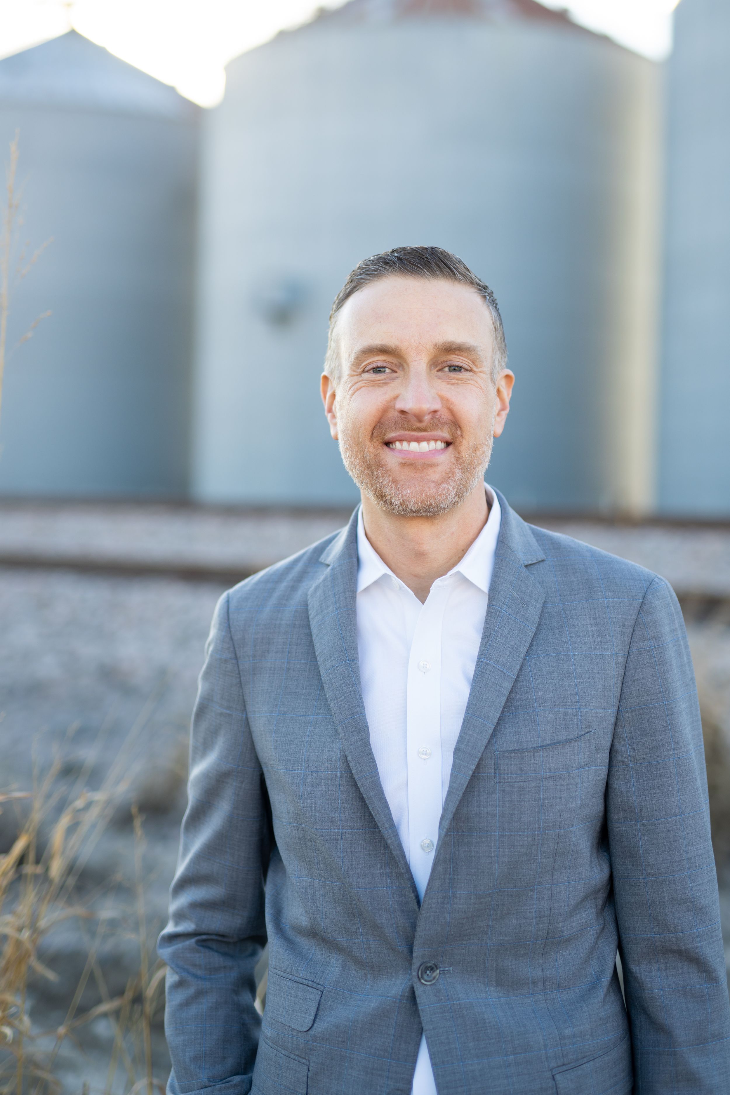 Professional portrait in a grey suit and white shirt standing in front of grain silos on a sunny day.