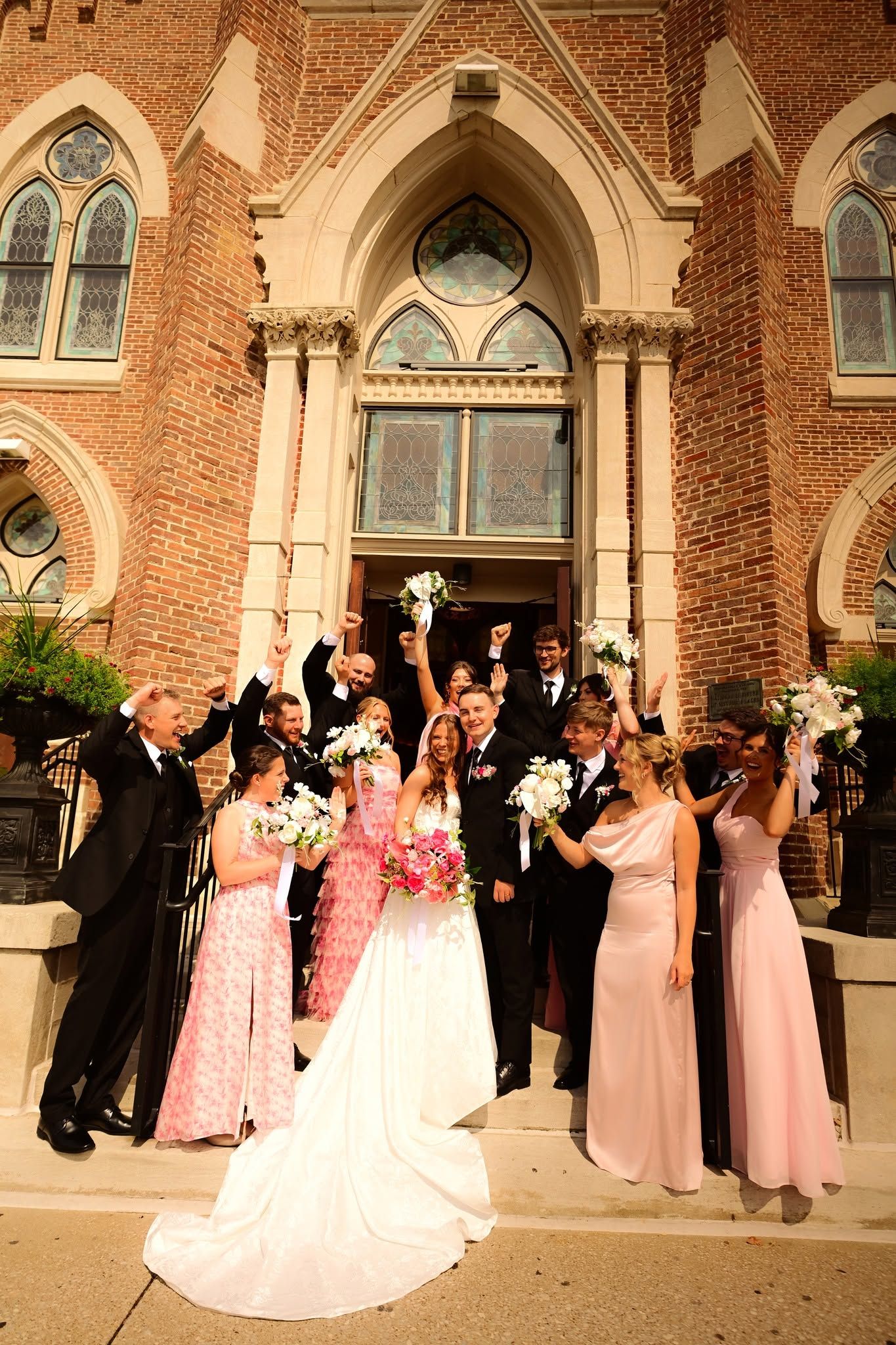 Wedding party celebrating on church steps, bride in white gown with pink bouquet, bridesmaids in pink dresses, groomsmen in black suits.