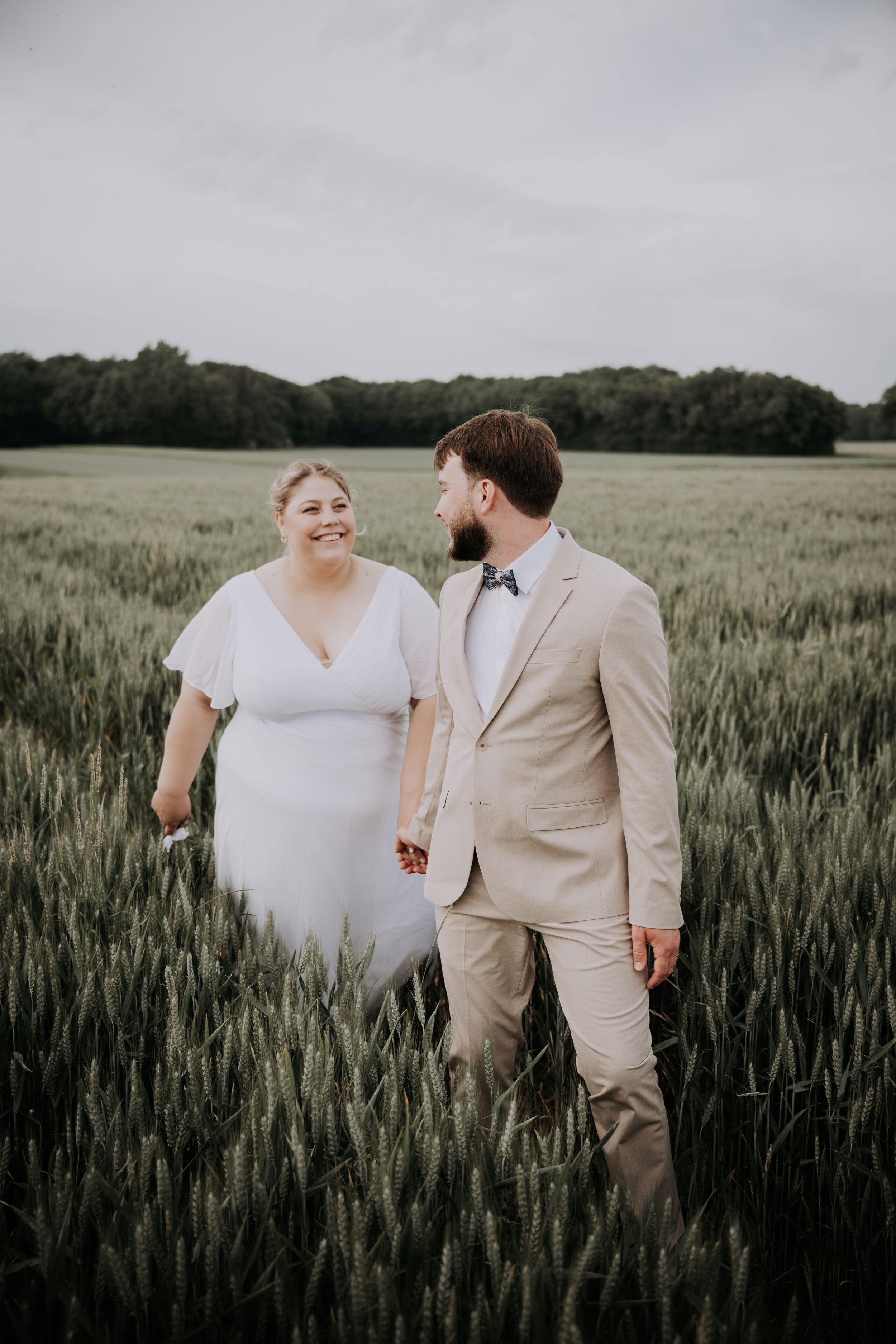 Bride and groom walking hand in hand through a lush green wheat field on their wedding day.