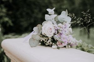 A romantic bridal bouquet of soft pink roses, lavender flowers, and eucalyptus rests on a white surface outdoors.