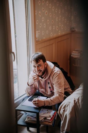 Smiling young man in pink hoodie sitting at a small desk by a window, resting his chin on his hand.