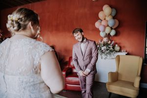 Groom in pink suit smiles at bride during first look moment, colorful balloons and flowers in background.
