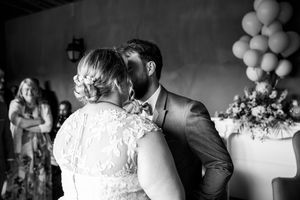 Bride and groom share first kiss at wedding ceremony in black and white photo with guests watching.