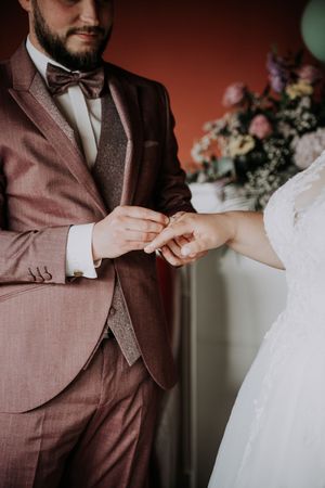 Groom in burgundy suit placing wedding ring on bride's finger during ceremony.