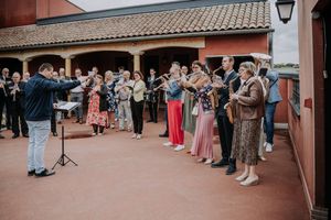 Conductor leads a choir group performing outdoors at a festive event on a terrace with red brick building.
