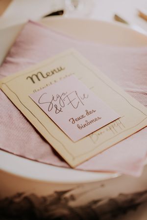Elegant wedding menu card with cursive script on pink linen napkin, featuring names Sego & Elie at a French reception.