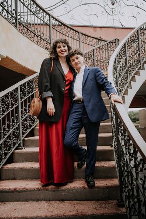 Couple posing on ornate spiral staircase, woman in red dress and black blazer, man in blue suit.