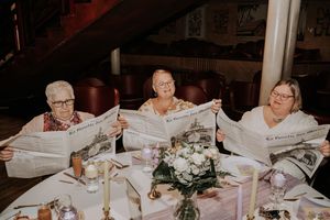 Three elderly women sitting at a decorated dinner table, each holding and reading a newspaper at a formal event.