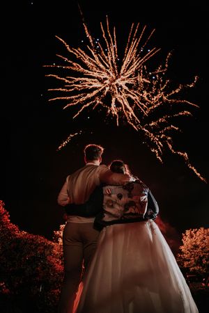 Bride and groom embrace while watching golden fireworks light up the night sky at their wedding celebration.