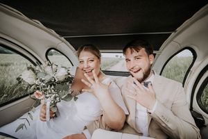 Newlyweds smiling and showing wedding rings inside a vintage car, bride holding a white bouquet.
