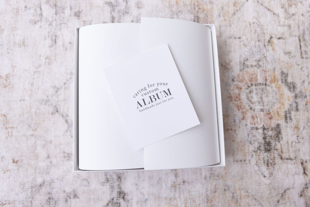 An open white gift box containing a photo album care card on a textured surface.
