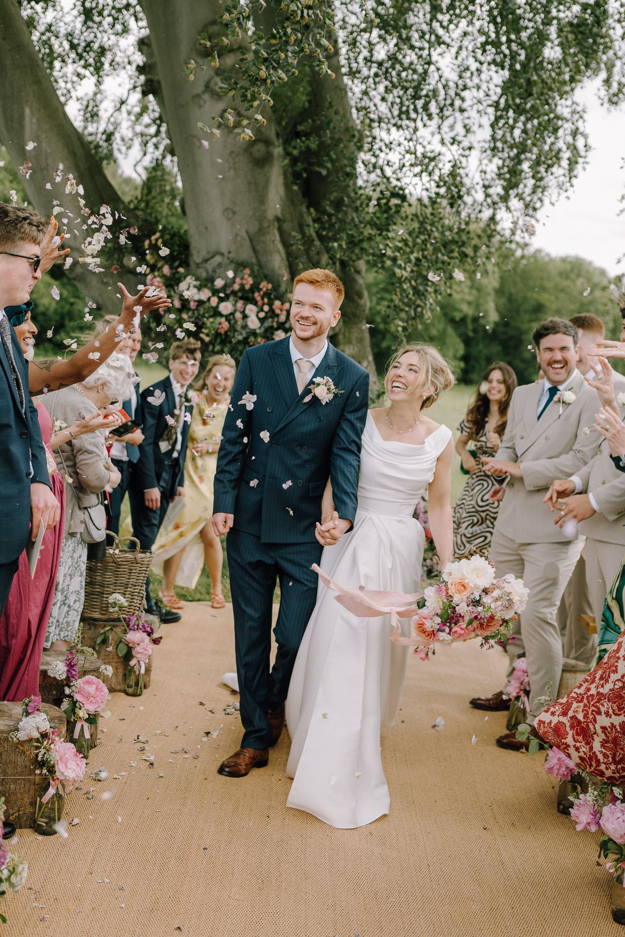 Joyful newlyweds walk through confetti shower surrounded by cheering guests outdoors at a garden wedding ceremony.