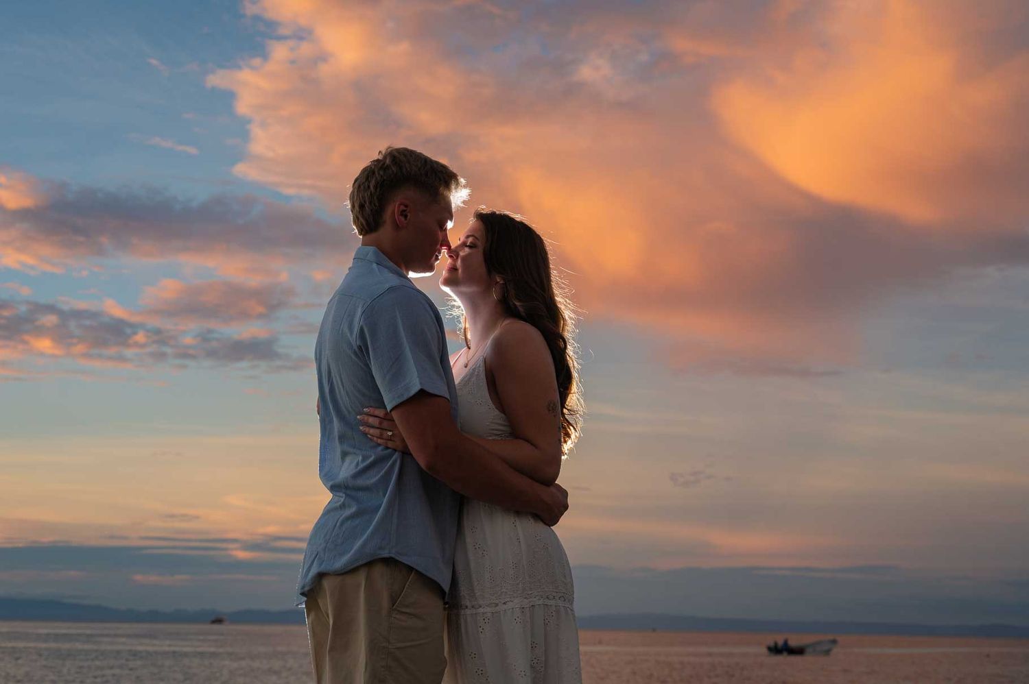 Couple embracing on beach at sunset, woman in white dress smiling as man kisses her head, ocean backdrop.