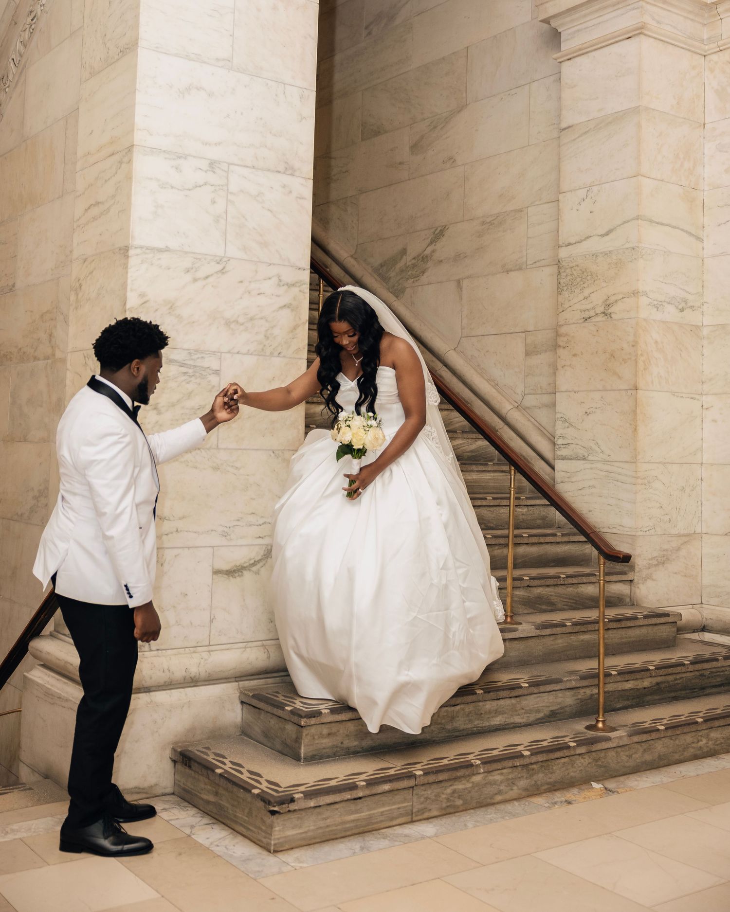 Groom in white tuxedo holding bride's hand on marble staircase.