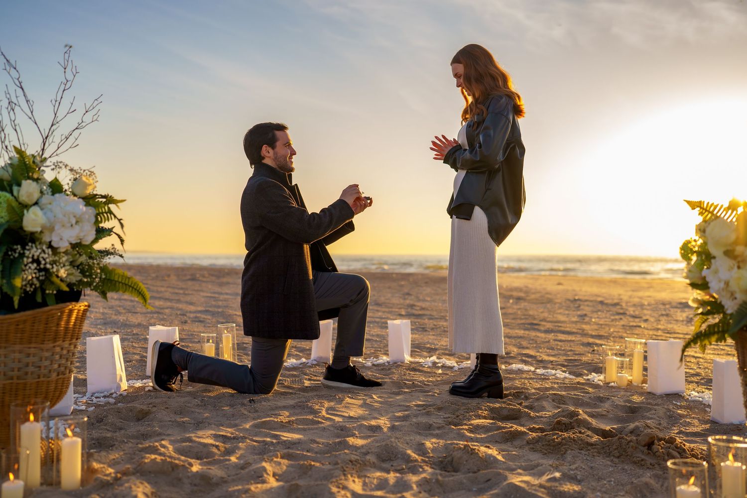 Man proposes to woman on a sandy beach at sunset, surrounded by candles and white flowers.