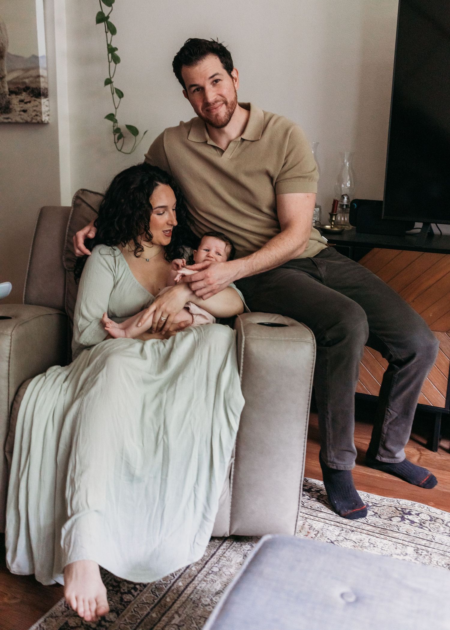 A couple sits on a sofa with their newborn baby in a cozy living room, sharing a tender family moment.