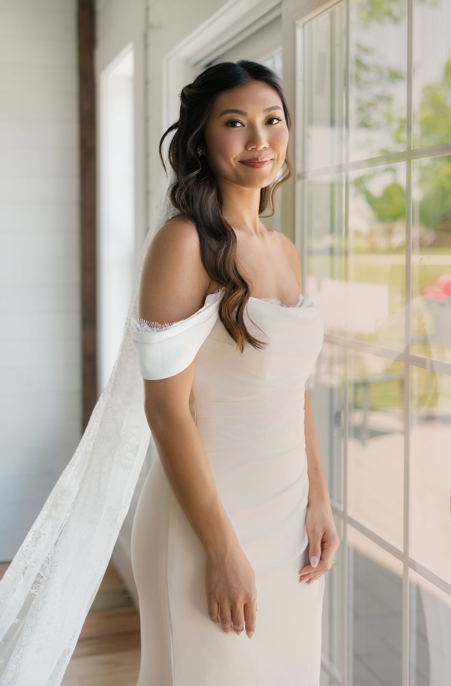 Bride in off-shoulder white gown standing by large window at williams farm pataskala, ohio.