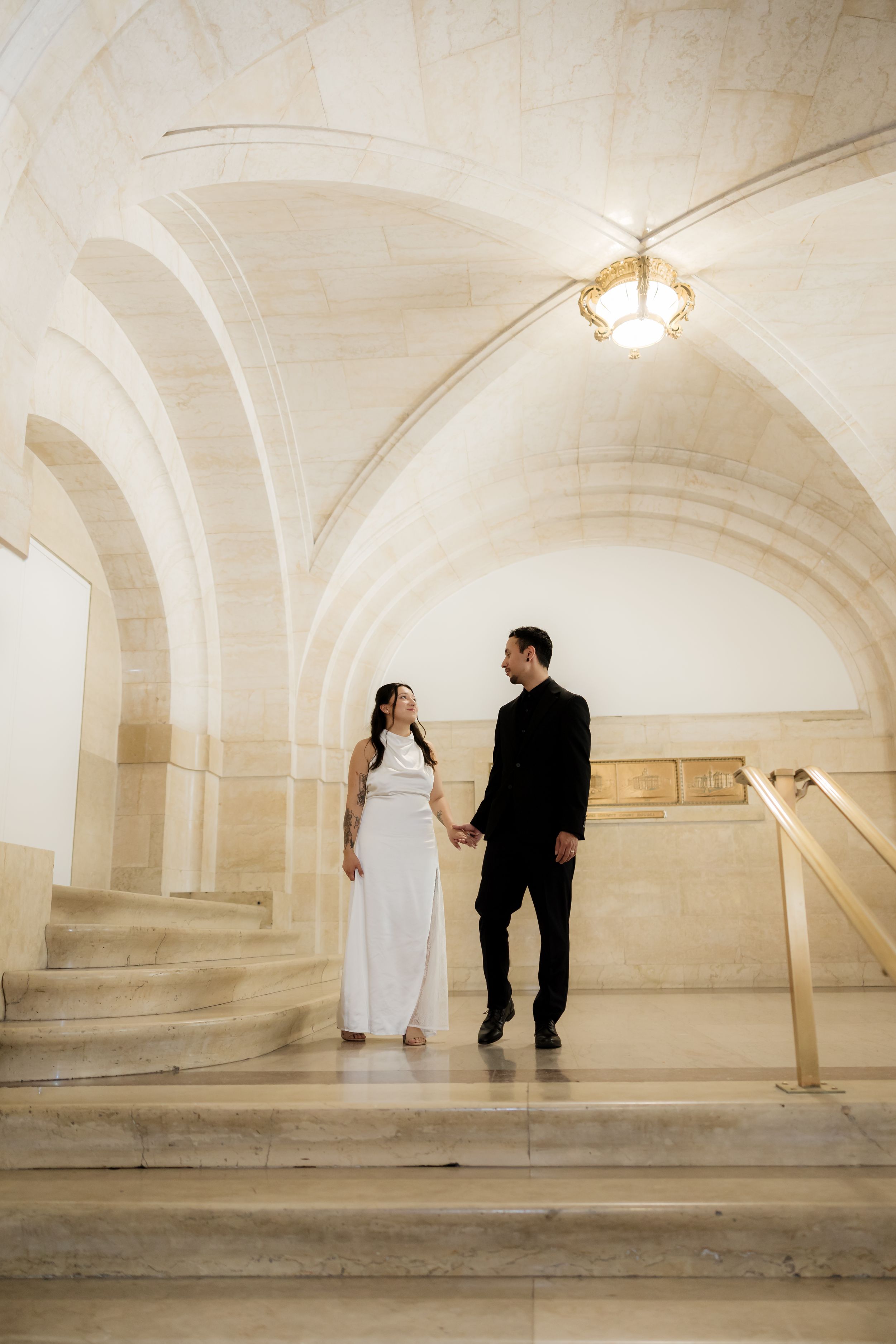 Couple in wedding attire holding hands on marble stairs under arched ceiling with elegant chandelier.
