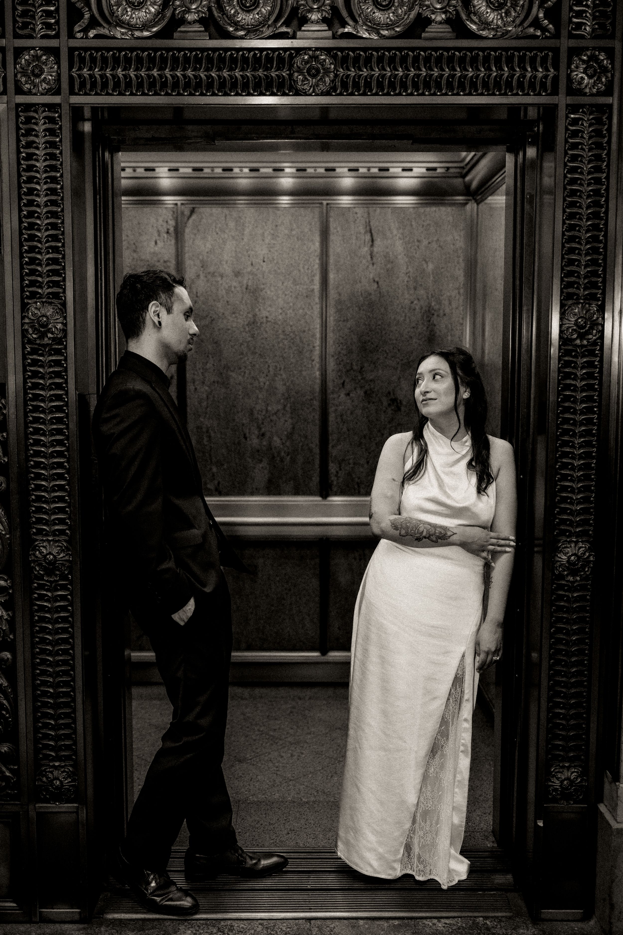 Black and white photo of a bride in white gown and groom in black suit standing in an ornate elevator.
