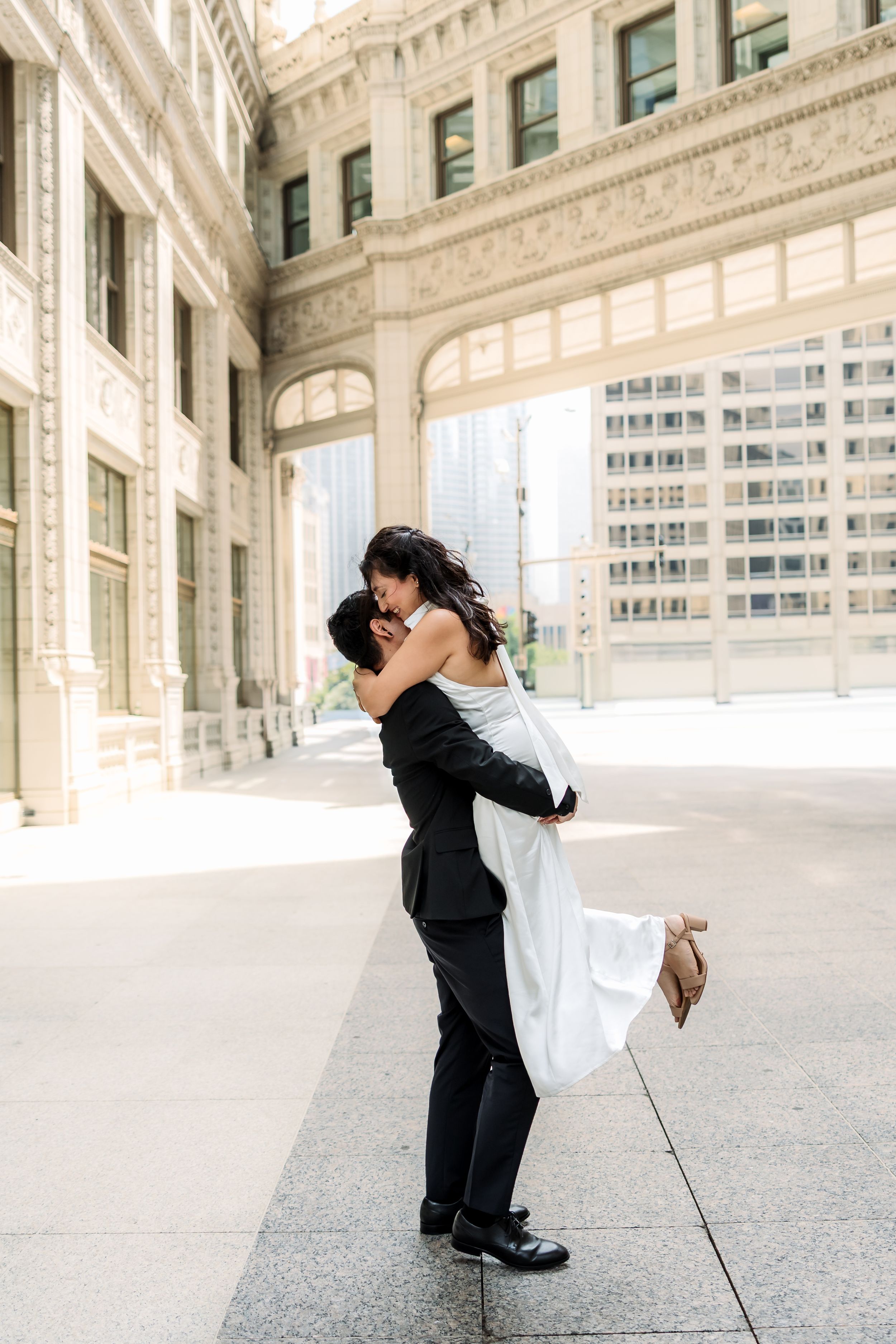 Groom lifting bride in white dress, sharing a romantic kiss outside a grand urban building on their wedding day.