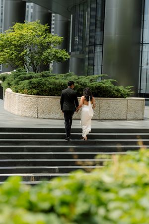 Newlywed couple holding hands walking up steps outside a modern glass building surrounded by greenery.