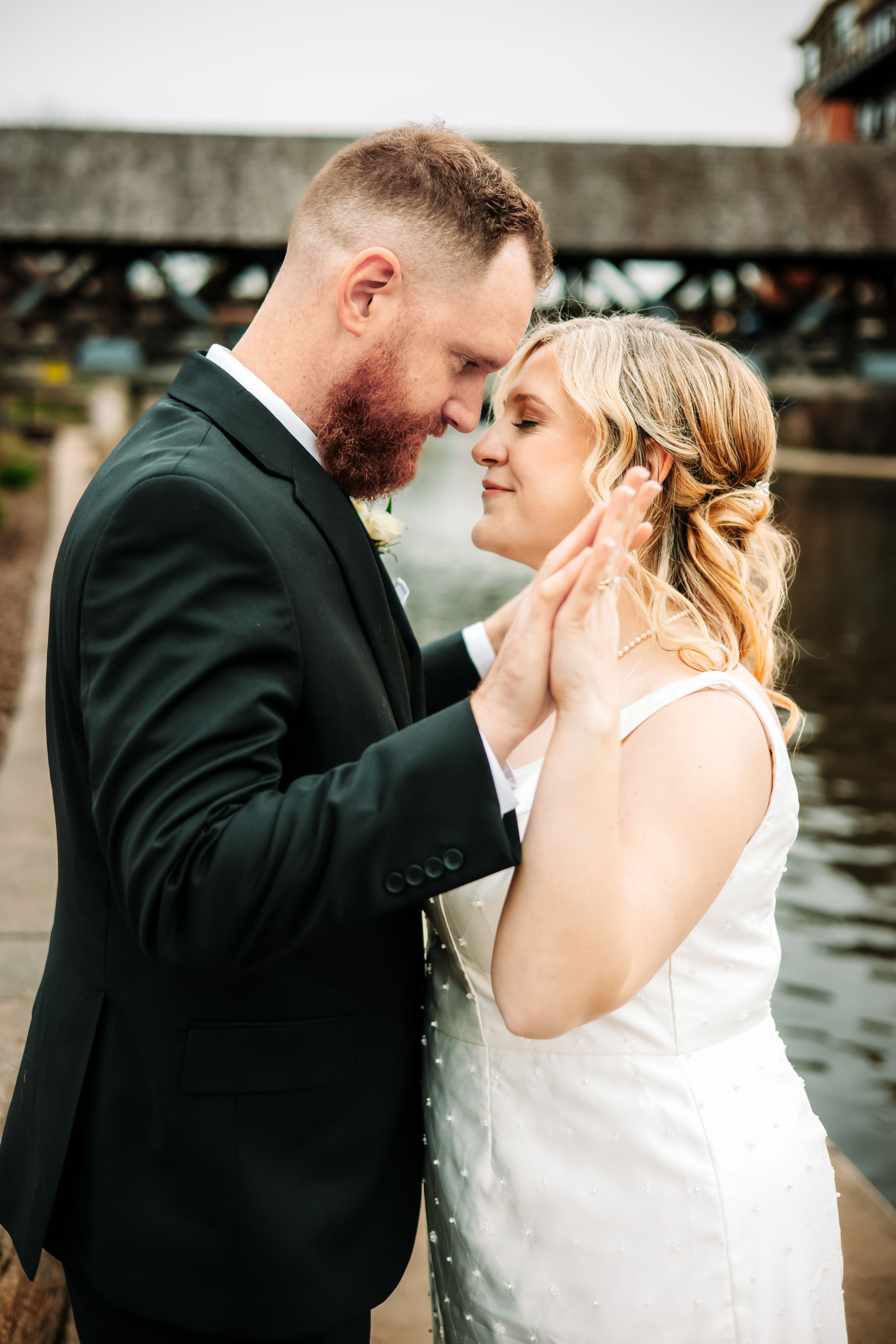 A groom in a black suit tenderly holds a bride's face near a waterway bridge in an intimate wedding portrait.